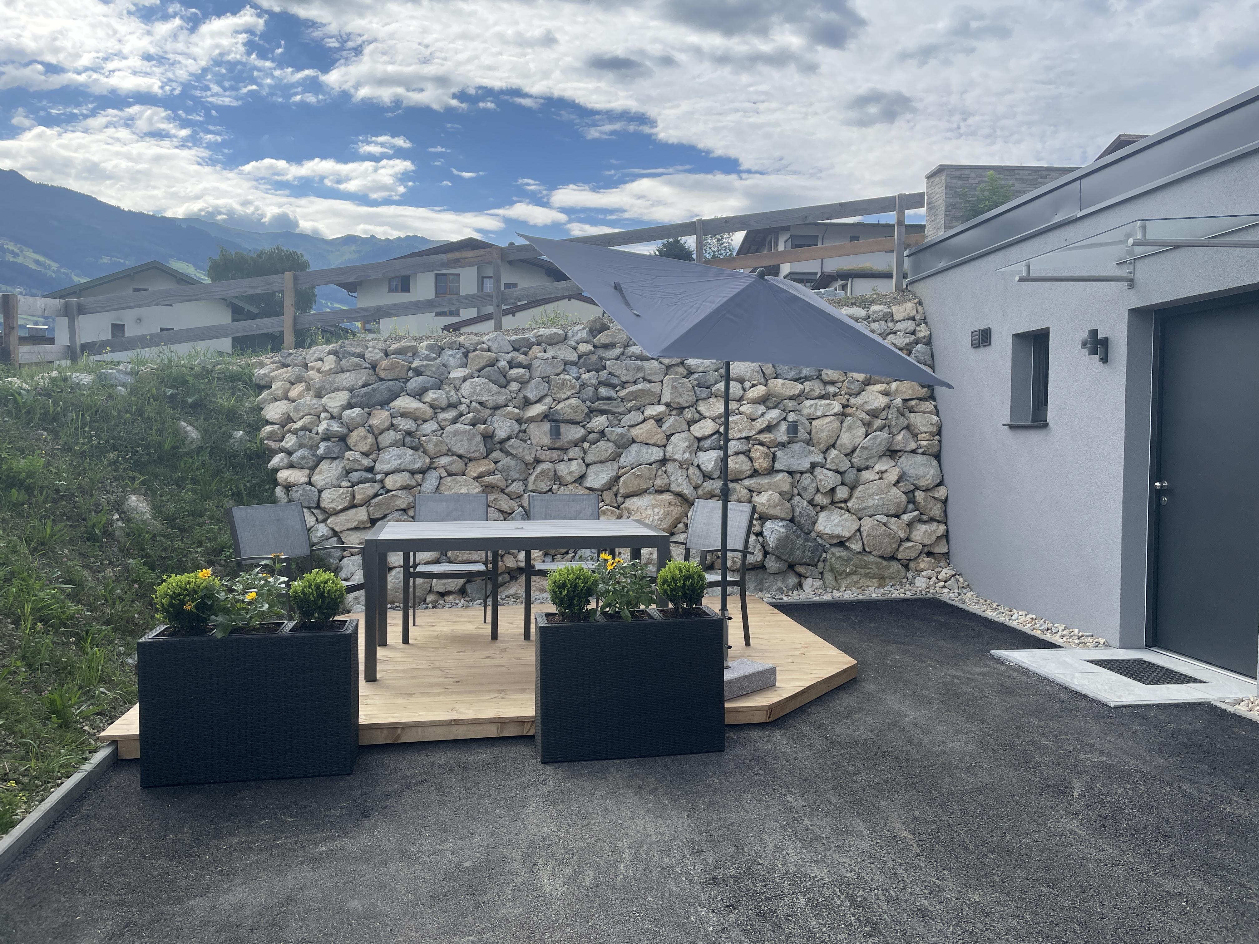 Ein moderner Außenbereich mit Terrasse, Stühlen, Sonnenschirm und Steinmauer vor einem Haus mit Bergen im Hintergrund.
