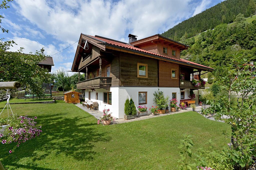 Häuser im Grünen mit Garten, Blumen und Hügel im Hintergrund, sonniger Tag, klare Wolken am Himmel.