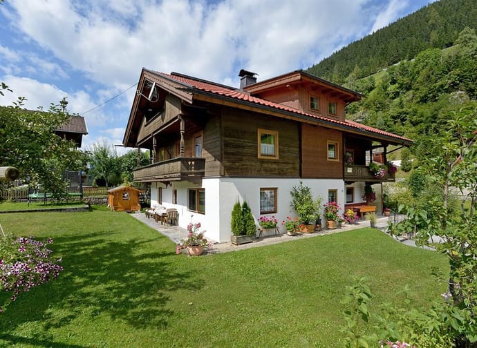 Häuser im Grünen mit Garten, Blumen und Hügel im Hintergrund, sonniger Tag, klare Wolken am Himmel.