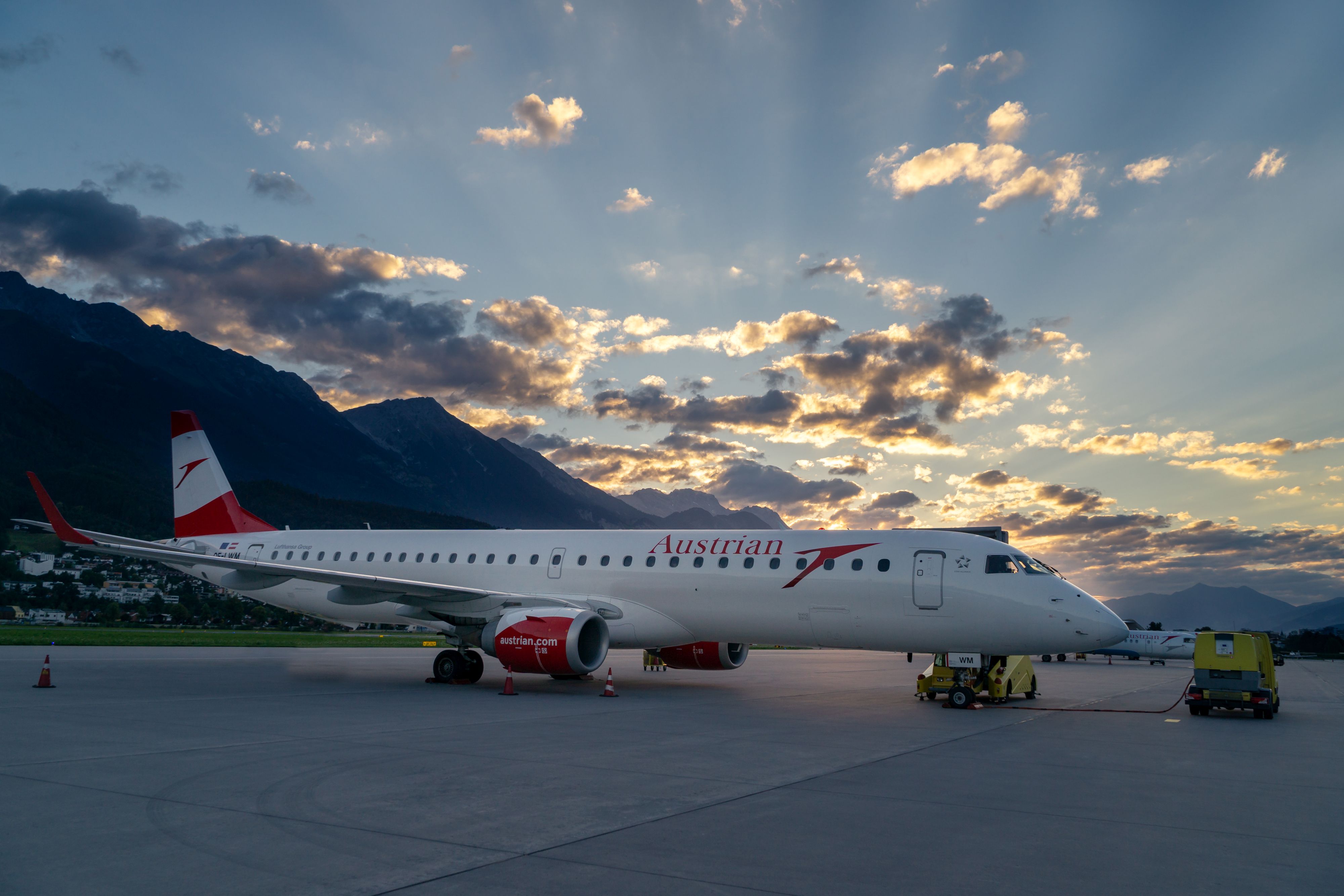 Austrian Airlines Flugzeug am Flughafen Innsbruck bei Sonnenuntergang. Das weiß-rote Flugzeug steht auf dem Vorfeld vor dramatischer Bergkulisse der Alpen mit bewölktem Abendhimmel.