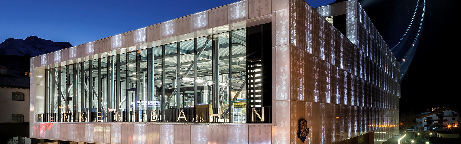 Exterior view of the Bründl Sports building at the Penken cable car base station in Mayrhofen. A modern glass facade reflects the evening sky, with the illuminated logo visible on the front and a wide entrance at street level.