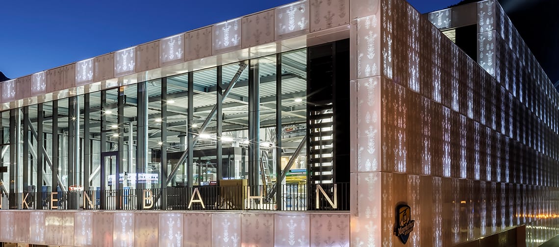 Außenansicht des modernen Bründl-Sports-Gebäudes an der Talstation der Penkenbahn in Mayrhofen. Große Glasfassade mit Beleuchtung, sichtbares Logo an der Front, breiter Eingangsbereich auf Straßenniveau.