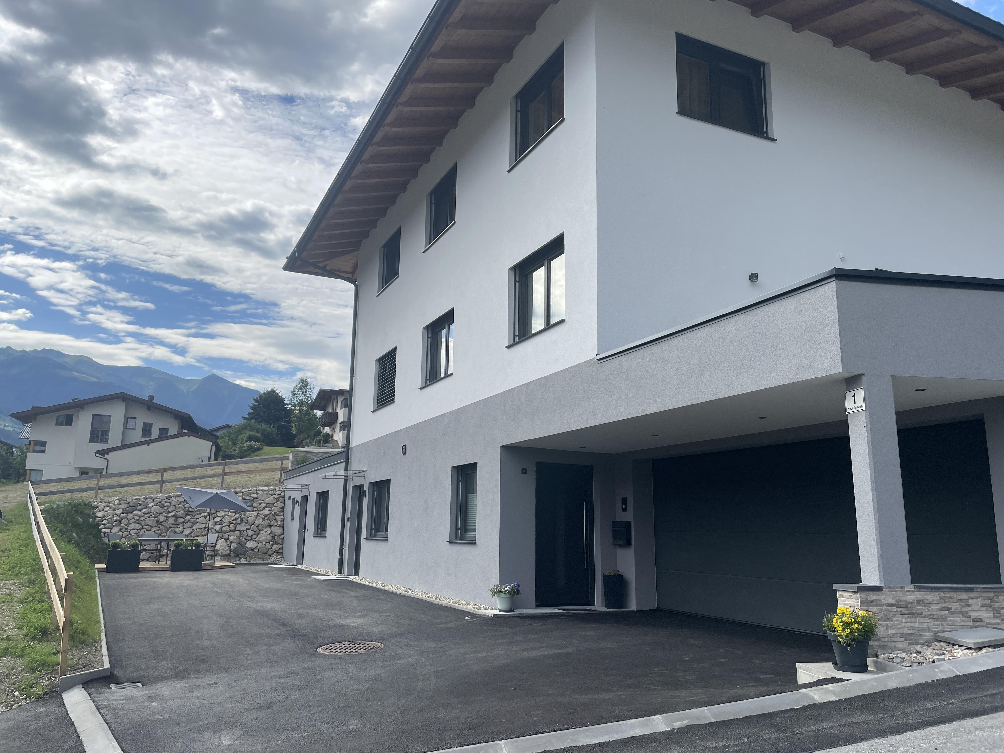 Modernes weißes Mehrfamilienhaus mit mehreren Fenstern, im Hintergrund Berge und bewölkter Himmel.