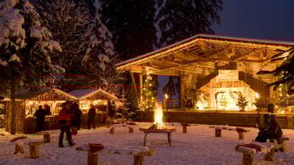 Das Bild zeigt einen stimmungsvollen Weihnachtsmarkt in einer winterlichen Waldkulisse mit einer beleuchteten Bühne, Holzbuden, einem Lagerfeuer und schneebedeckten Sitzbänken.