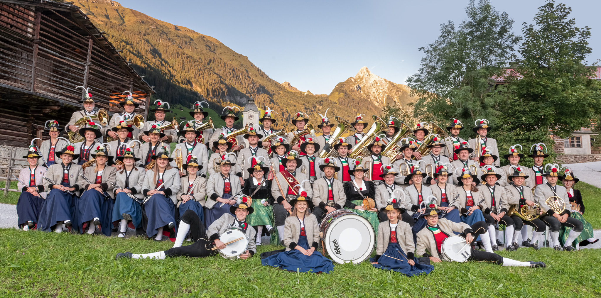 The brass band poses in traditional attire on a meadow with an impressive mountain backdrop for a group photo.