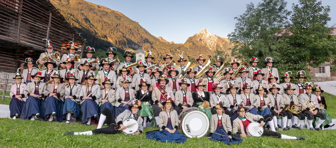The brass band poses in traditional attire on a meadow with an impressive mountain backdrop for a group photo.