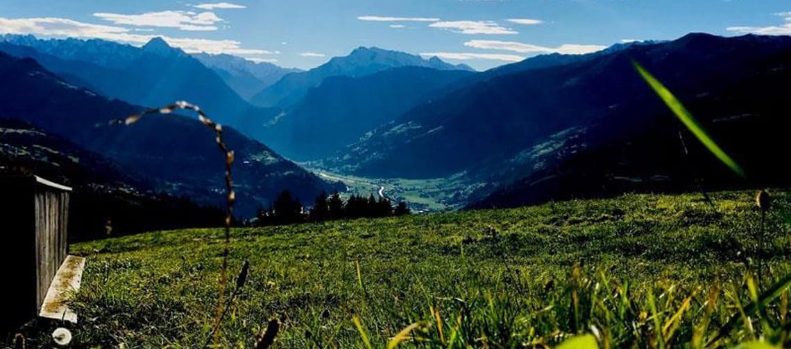 Berglandschaft mit grünen Wiesen, Bergen im Hintergrund und sonnigem Himmel, Sonnenstrahlen sichtbar.