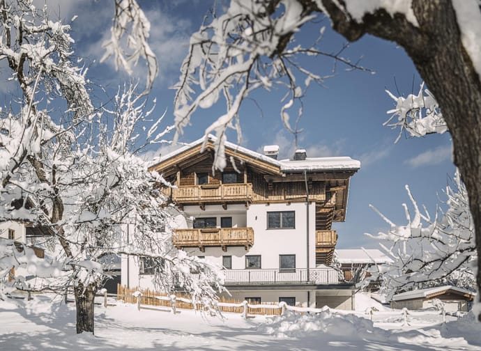 Ein modernes Chalet im Schnee, umgeben von verschneiten Bäumen unter blauem Himmel.