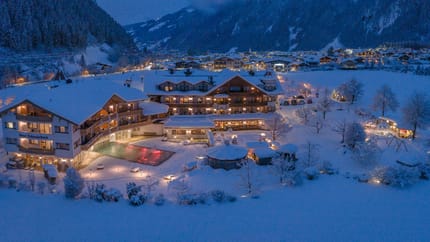 Ein verschneites Hotel in den Bergen bei Nacht, mit beleuchteten Fenstern und einem beheizten Pool im Vordergrund.