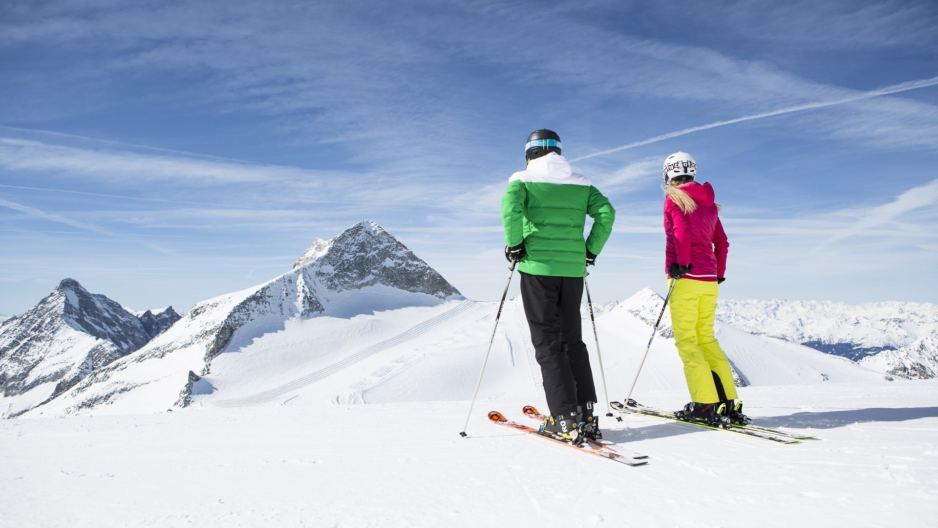 Zwei Skifahrer:innen stehen am schneebedeckten Hintertuxer Gletscher. Vor ihnen ragen verschneite Gipfel in den blauen Himmel. Sie blicken in die Weite der imposanten Berglandschaft.