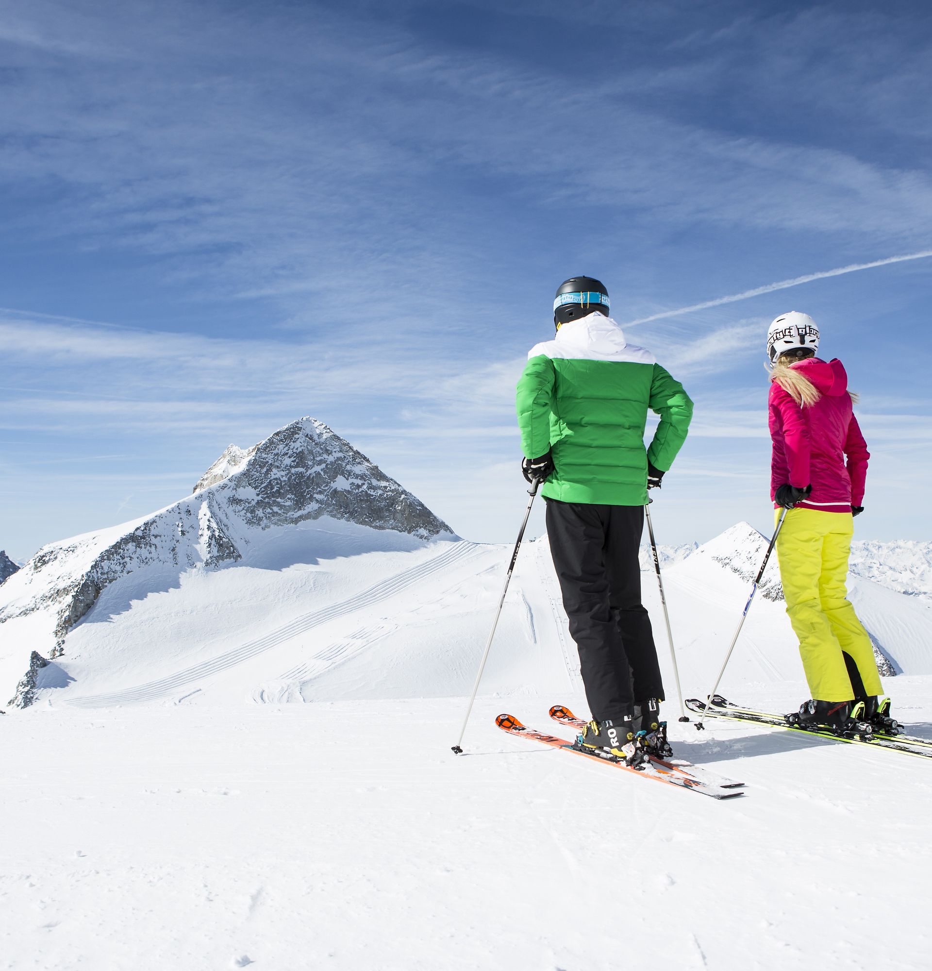 Two skiers stand on the snow-covered Hintertux Glacier. In front of them, snow-covered peaks rise into the blue sky. They gaze out into the vastness of the imposing mountain landscape.
