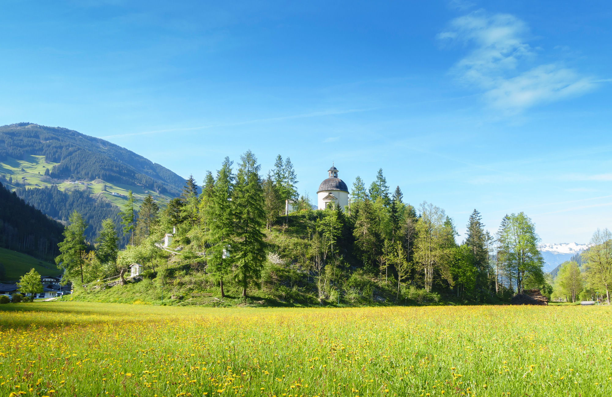 View of the forested Burgschrofen hill with chapel on the Schwendau Trail in Tyrol, surrounded by meadows and mountains, Station 3 “Burgschrofen,” photographed by Paul Wechselberger.