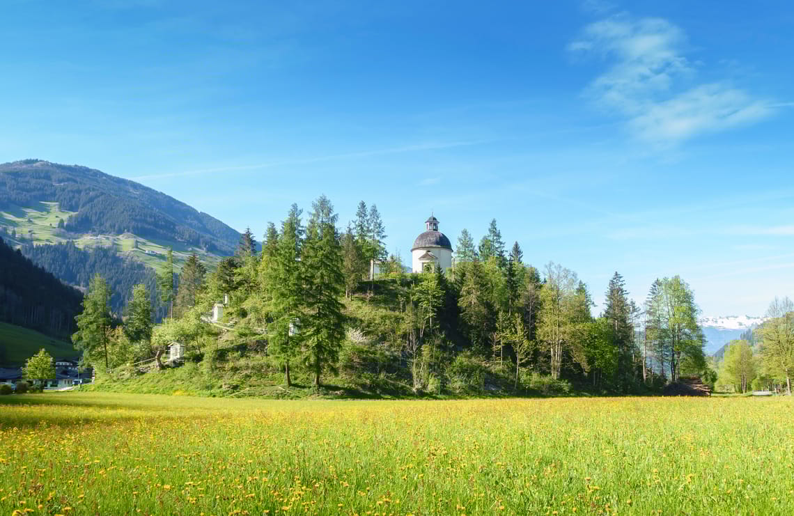Schwendauer Pfad - Burgschrofen Blick auf den bewaldeten Hügel Burgschrofen mit Kapelle am Schwendauer Pfad in Tirol, umgeben von Wiesen und Bergen, Station 3 „Burgschrofen“, fotografiert von Paul Wechselberger.