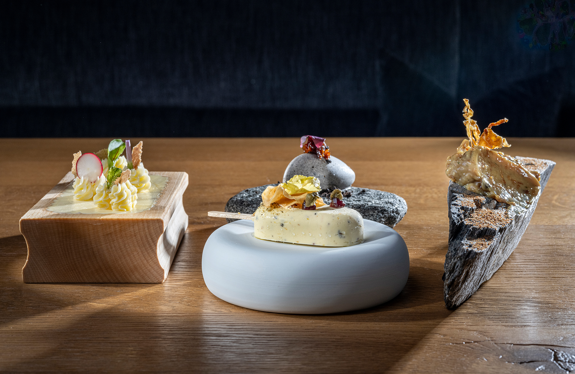 Drei kunstvoll angerichtete Gourmet-Häppchen auf einem Holztisch. Links auf einem Holzblock, in der Mitte als Eis am Stiel auf einem runden Stein und rechts auf einem Rindenstück, modern inszeniert im Hotel ZillergrundRock.