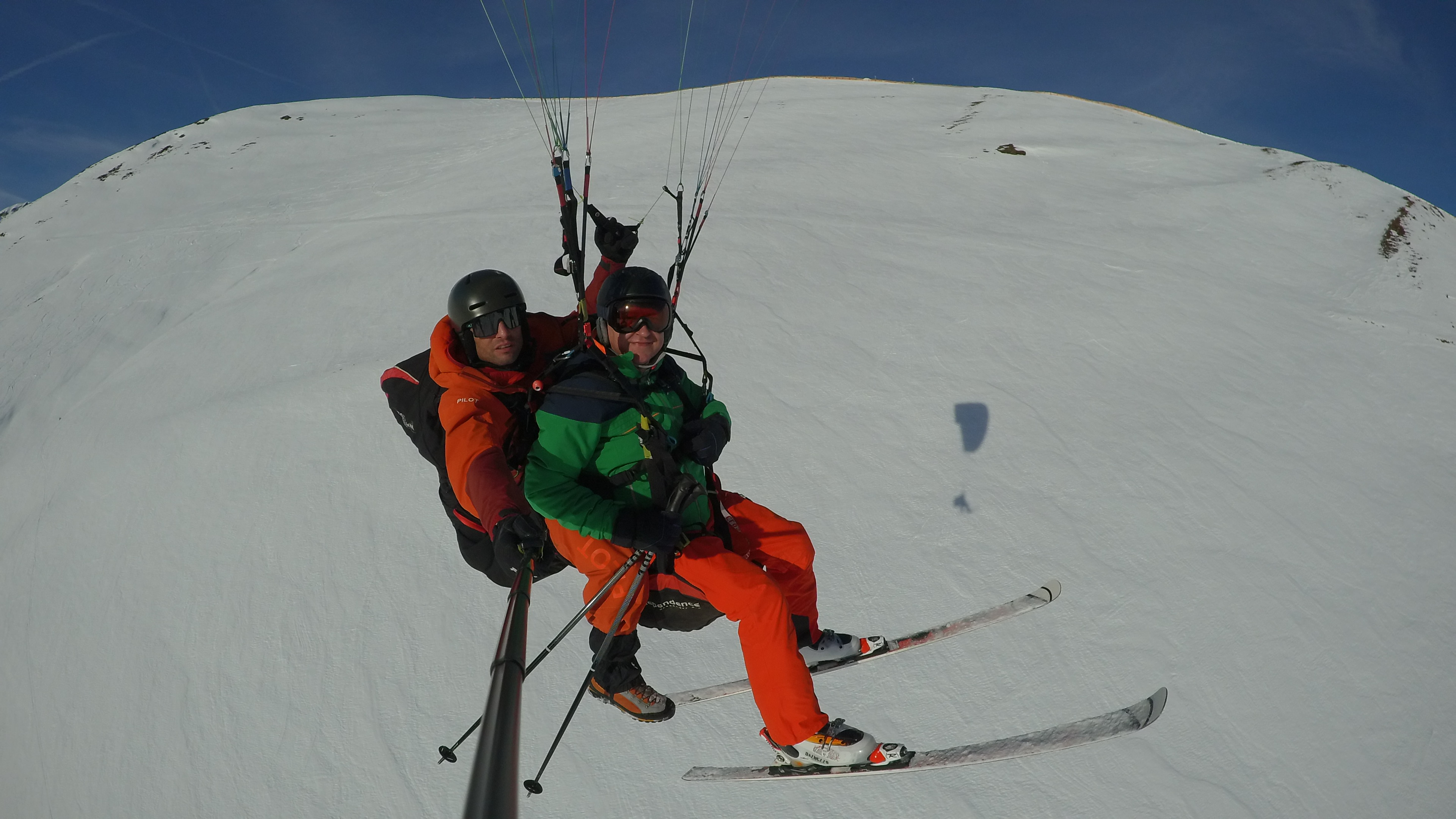 Zwei Personen in einem Paraglider über der Skipiste. Eine Person ist mit Ski auf dem Paraglider.