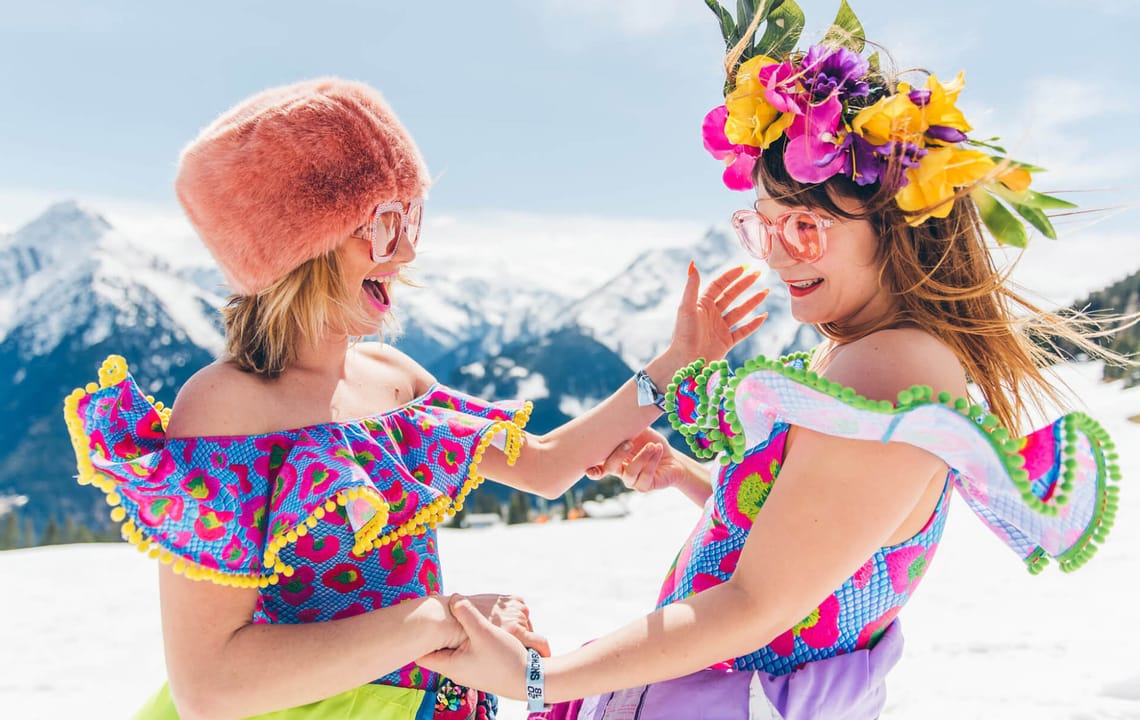 Snowbombing in Mayrhofen Snowbombing Festival Mayrhofen: Two women in colorful costumes dance and laugh in the snow with Alpine mountains in the background, one wearing a flower crown, the other a fur hat.