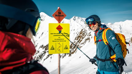Skier in blue gear with helmet and goggles, snowy mountains, near ski route sign.