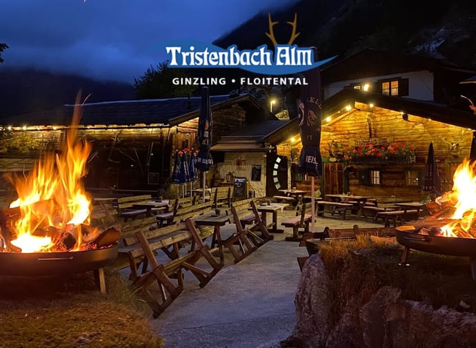Vor der rustikalen Holzhütte befinden sich offene Feuerstellen, umgeben von Holzbänken und Tischen. Warmes Licht, alpine Architektur und die Bergkulisse im Hintergrund schaffen eine gemütliche, urige Atmosphäre.