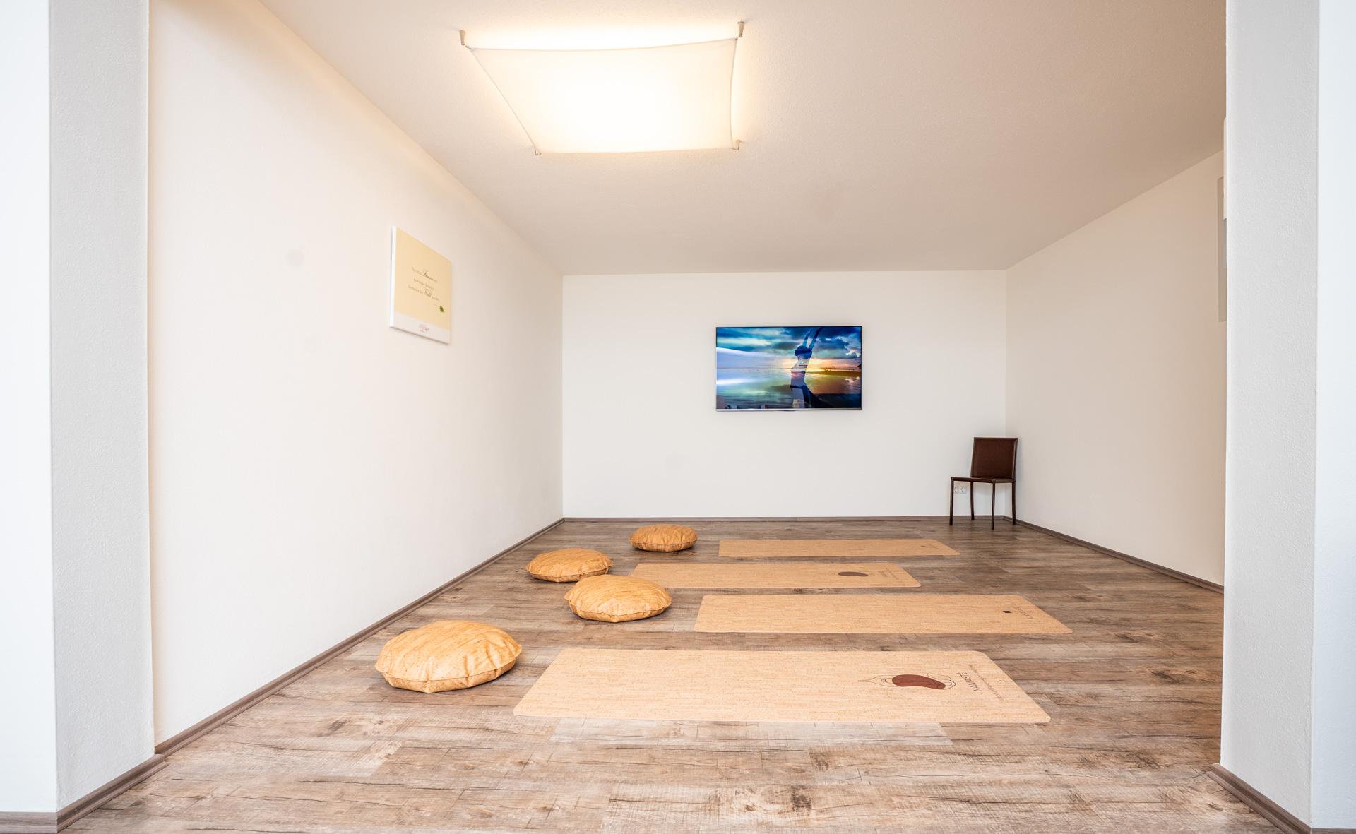 Ein leerer Raum mit weißen Wänden, Holzfußboden, Sitzkissen, Yogamatten, Wandbild und Stuhl.