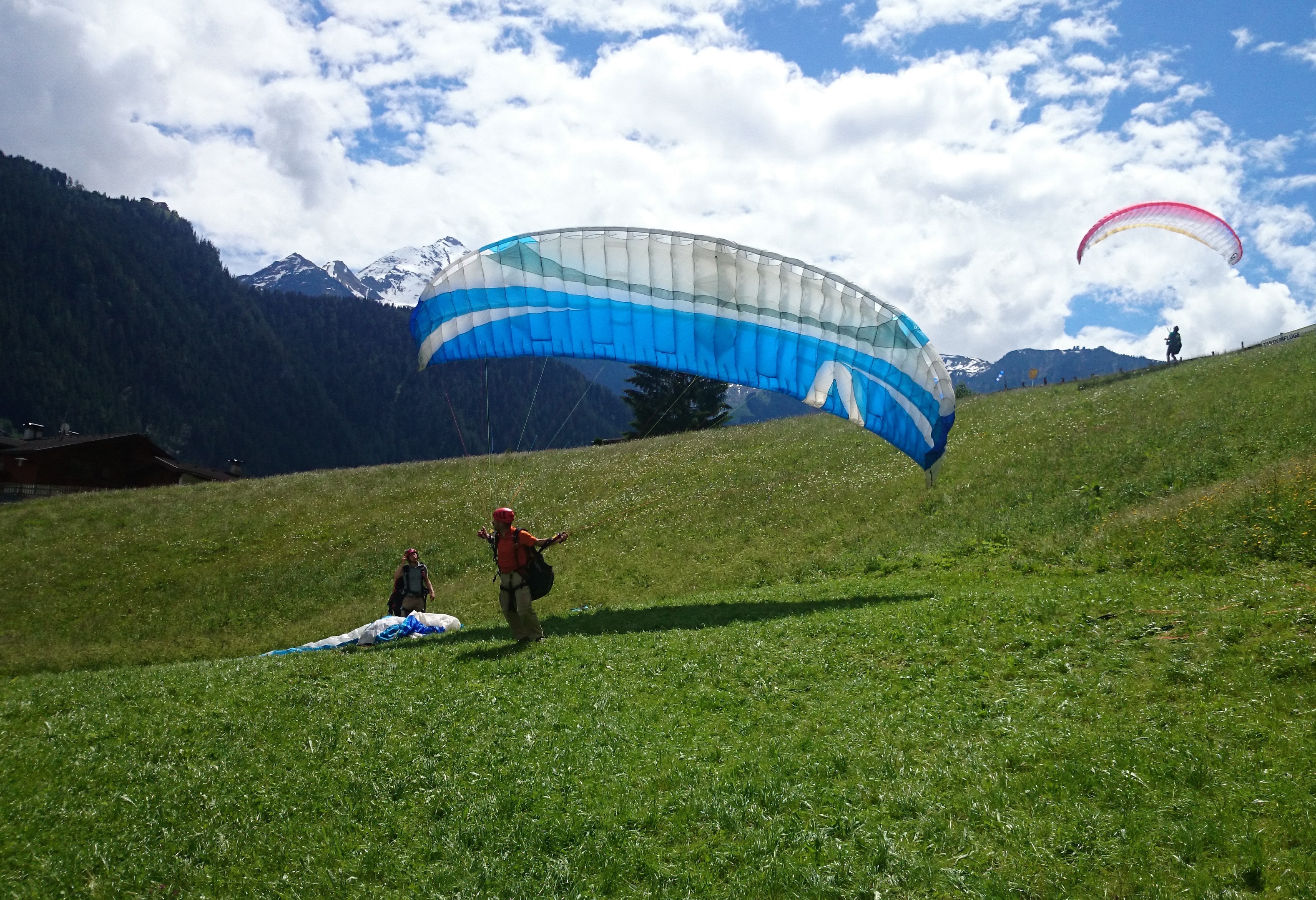 A paragliding student takes off with a blue and white canopy on a green meadow in the holiday region of Mayrhofen-Hippach, Zillertal, Tyrol. Another paraglider is in the air.