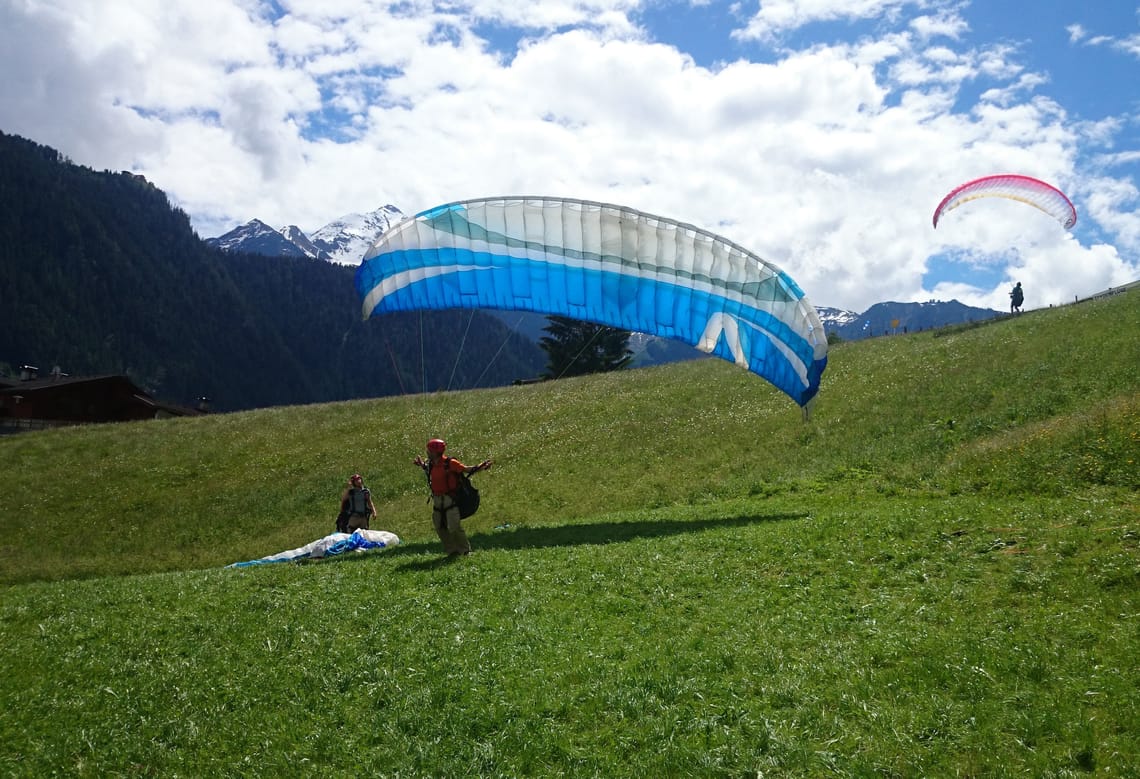 Paragleitkurse in Mayrhofen Ein Paragleitschüler startet mit einem blau-weißen Schirm auf einer grünen Wiese in der Ferienregion Mayrhofen-Hippach im Zillertal, Tirol. Ein weiterer Paragleiter ist in der Luft.