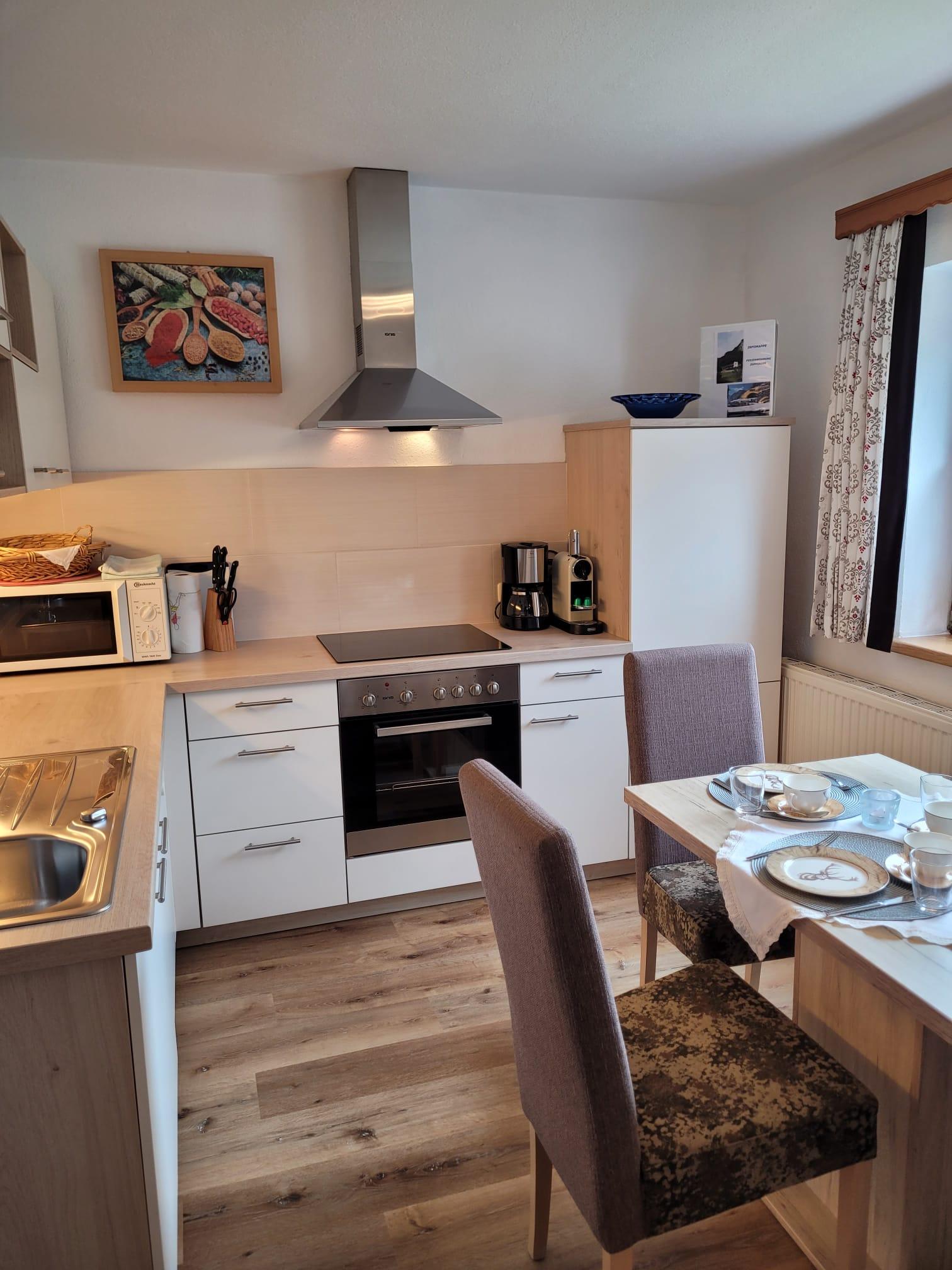 Küche mit Herd, Mikrowelle, Kaffeemaschine, Esstisch mit Besteck und Stühlen, Fenster mit Vorhängen.