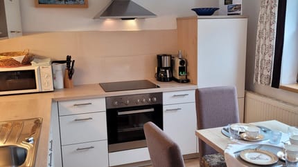 Küche mit Herd, Mikrowelle, Kaffeemaschine, Esstisch mit Besteck und Stühlen, Fenster mit Vorhängen.