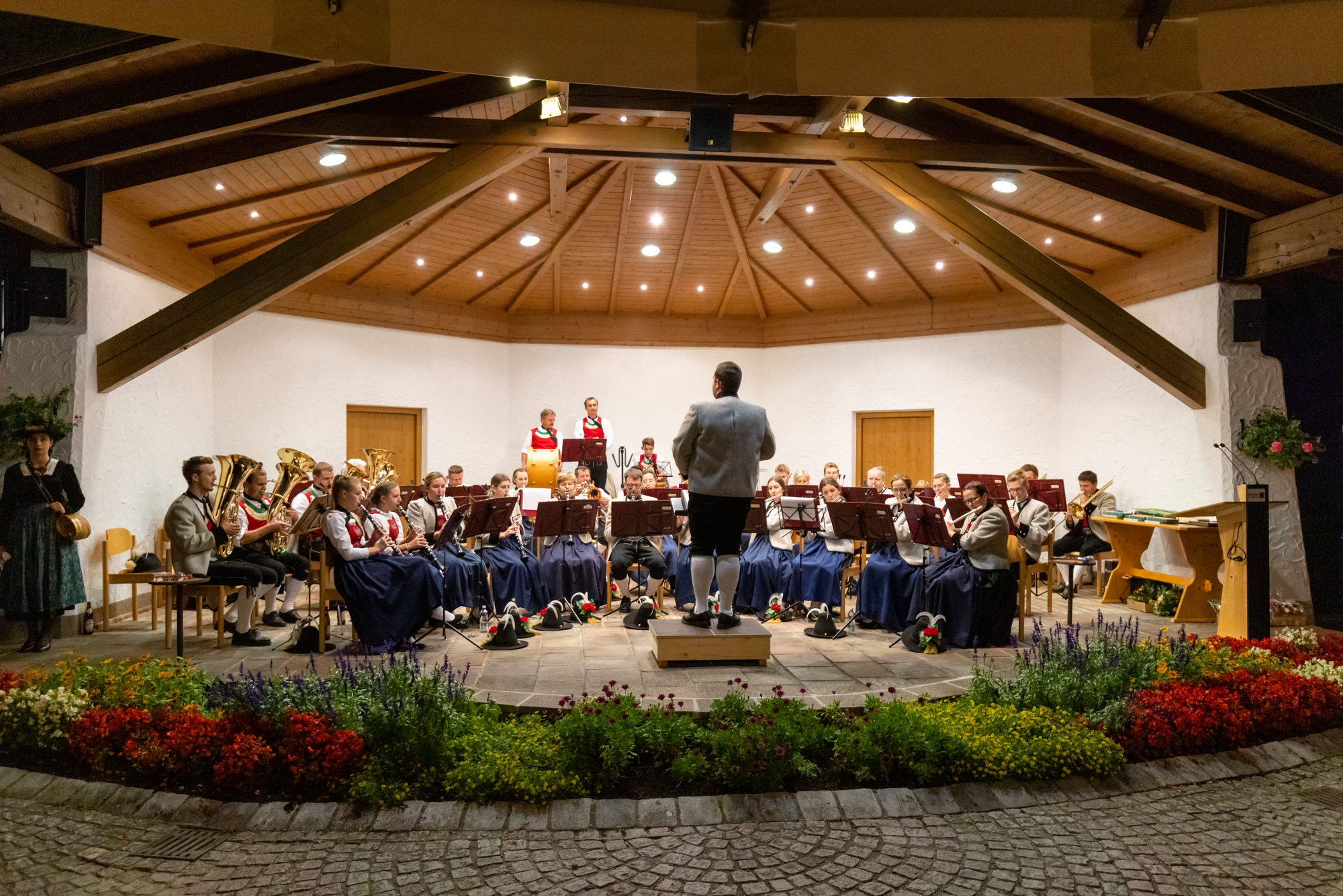 Das Bild zeigt eine Blaskapelle in traditioneller Tracht, die in einem überdachten Pavillon ein Konzert gibt. Der Dirigent steht auf einem kleinen Podest und leitet das Orchester.