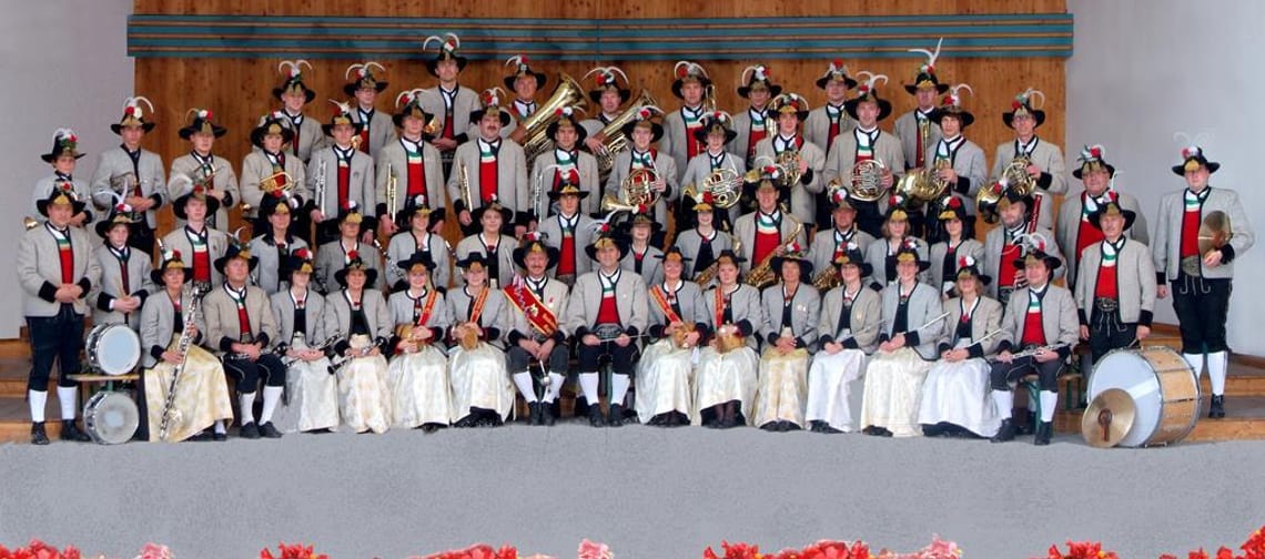 The image shows a large brass band in traditional attire posing for a group photo on a stage. The musicians wear festive hats adorned with feathers and play various brass instruments as well as drums.