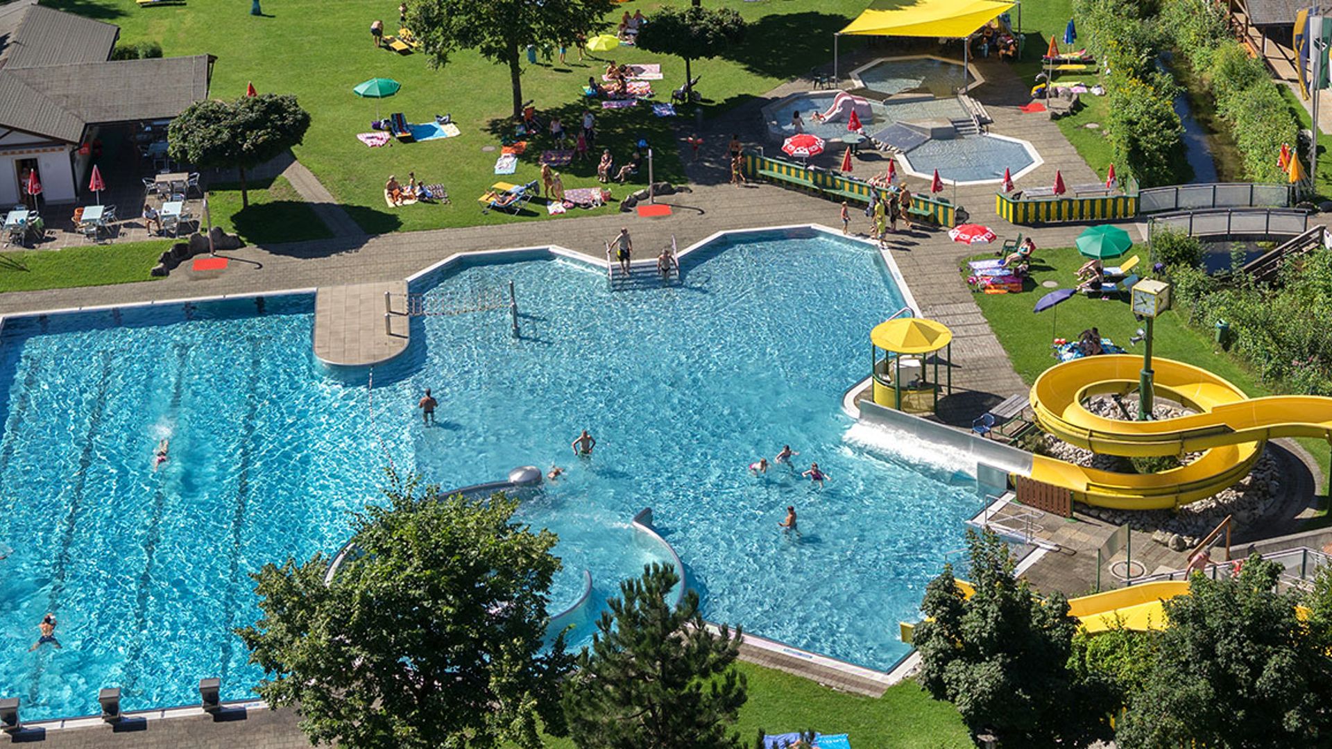 Aerial view of the outdoor pool at Summerworld Hippach in Zillertal with leisure pool, sports pool, slides, kids’ area, and large sunbathing lawns – perfect for families and swimmers.