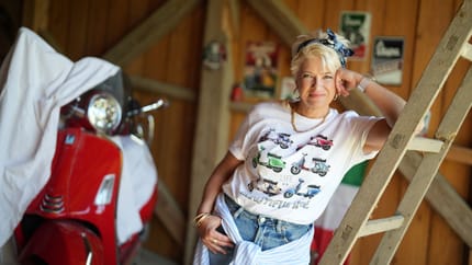 The singer Monique is standing in front of a red moped and leaning against a ladder.