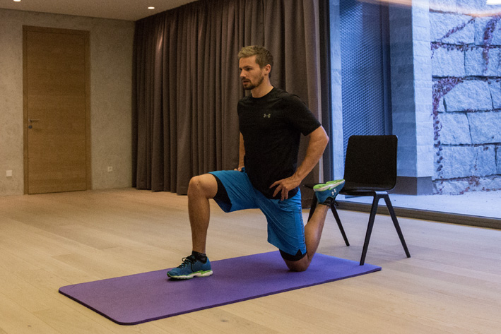 Mann führt Ausfallschritt mit hinterem Bein auf Stuhl in Trainingsraum in Mayrhofen aus. Christoph Ebenbichler zeigt Übung zur Stärkung von Beinen und Hüfte – ideal für Skifitness und Wintertraining.