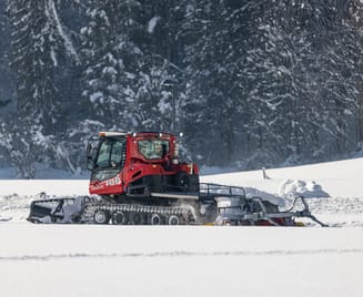 A red snow groomer preparing the cross-country trail in Mayrhofen-Hippach, surrounded by snowy fields and a winter forest