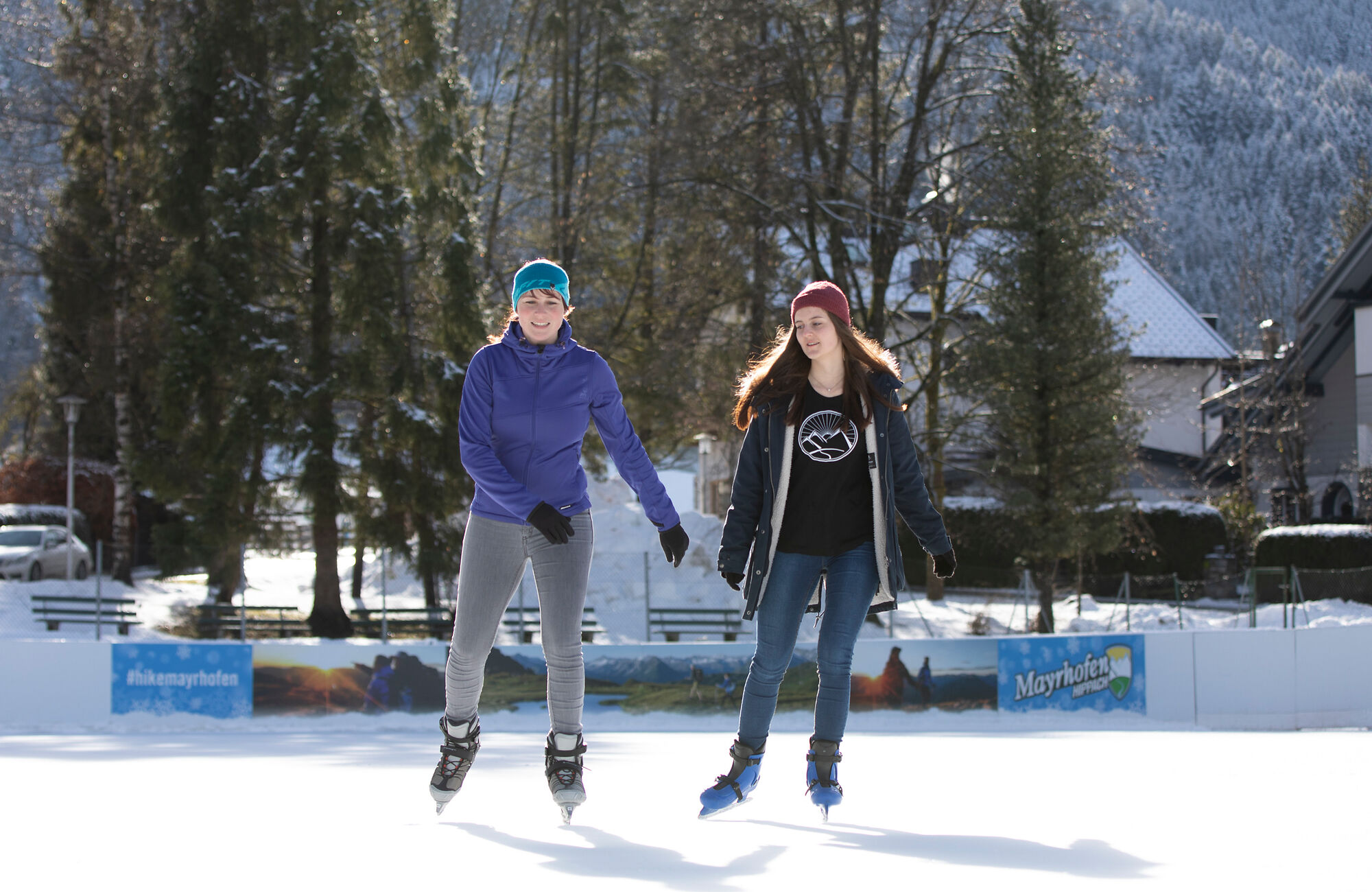 Zwei junge Menschen haben Spaß beim Eislaufen auf dem Eislaufplatz Mayrhofen im Zillertal. Vor winterlicher Bergkulisse genießen sie die Bewegung auf dem Eis unter blauem Himmel.