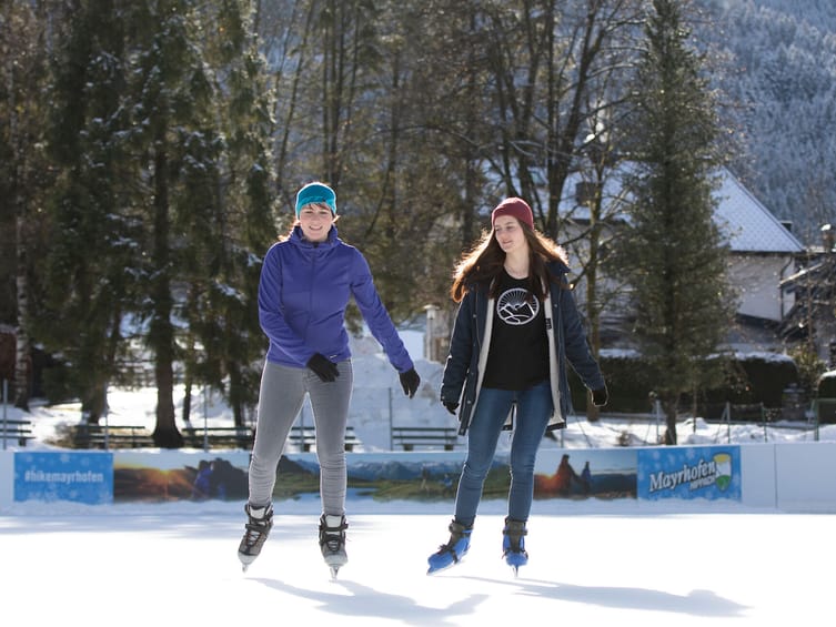 Zwei junge Menschen haben Spaß beim Eislaufen auf dem Eislaufplatz Mayrhofen im Zillertal. Vor winterlicher Bergkulisse genießen sie die Bewegung auf dem Eis unter blauem Himmel.