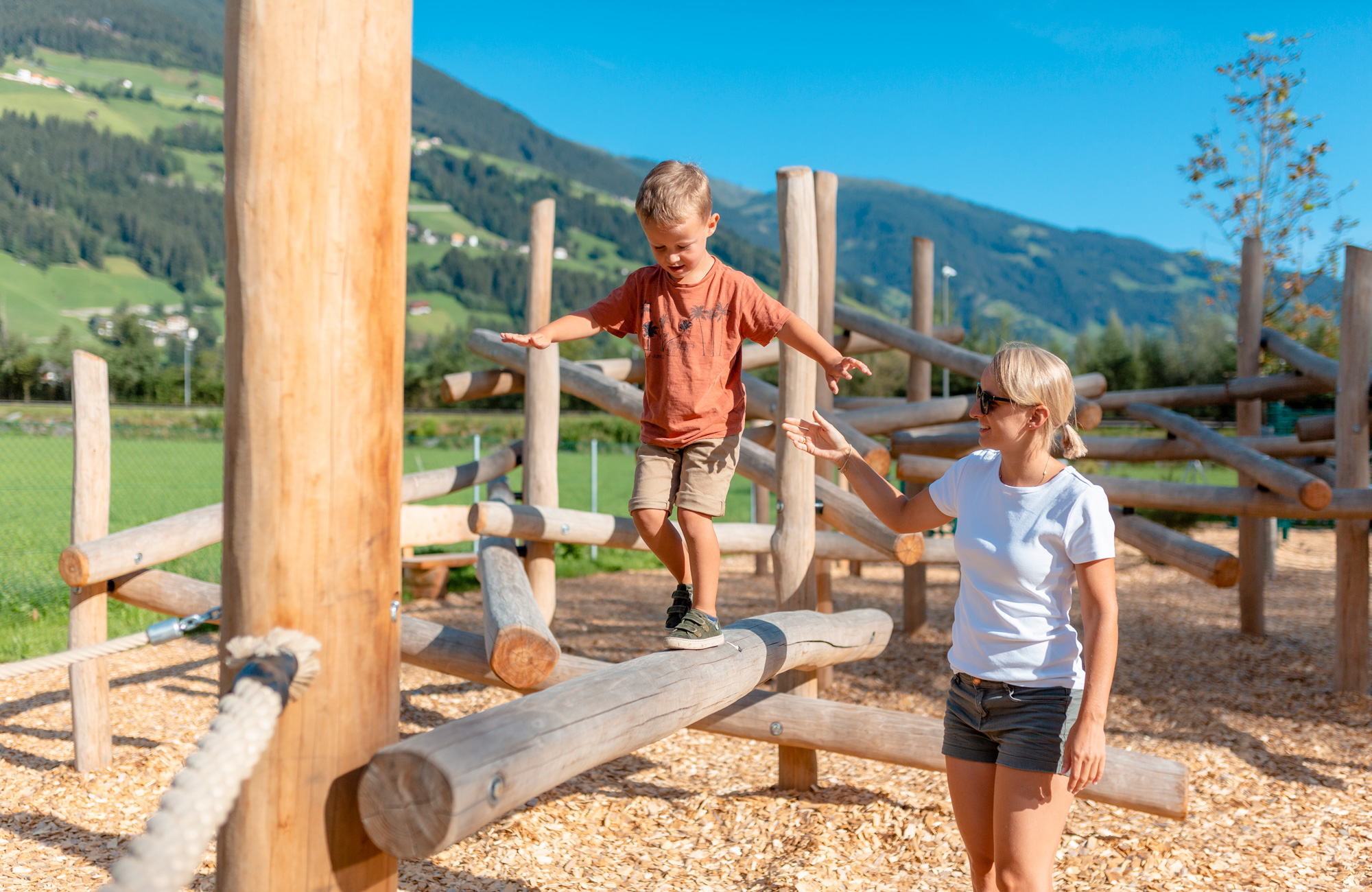 Children playing on a wooden balance course in Auenland Sidan on the Schwendau Trail in Tyrol, surrounded by green meadows and mountain scenery, Station 2 “Auenland Sidan,” photographed by Thomas Eberharter Photography.