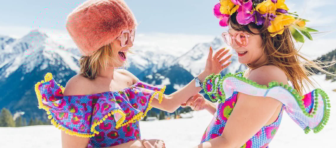 The imaThe image shows two joyful women in colorful, festive outfits with summery accessories, dancing and laughing in a snow-covered mountain landscape.
