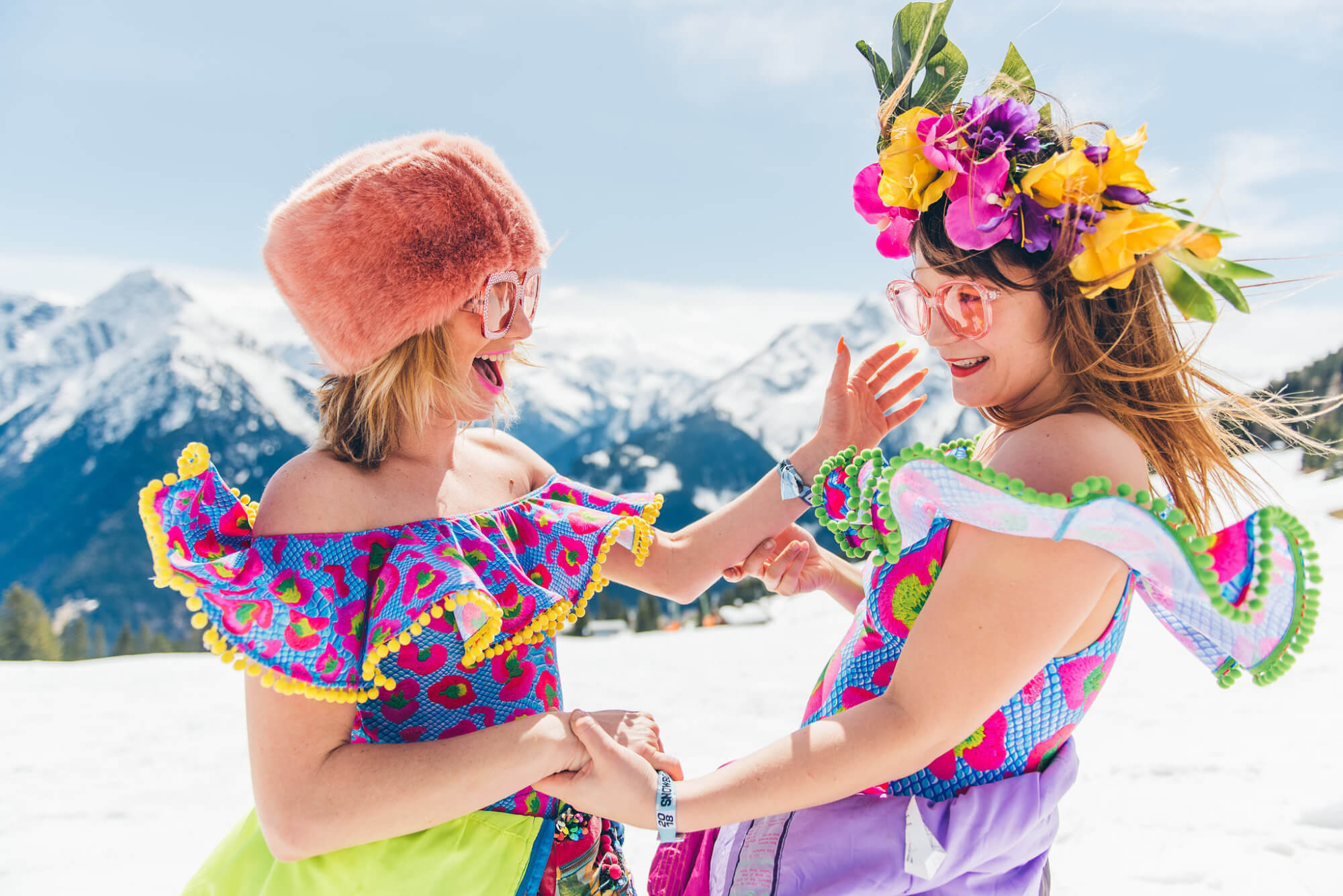 The imaThe image shows two joyful women in colorful, festive outfits with summery accessories, dancing and laughing in a snow-covered mountain landscape.