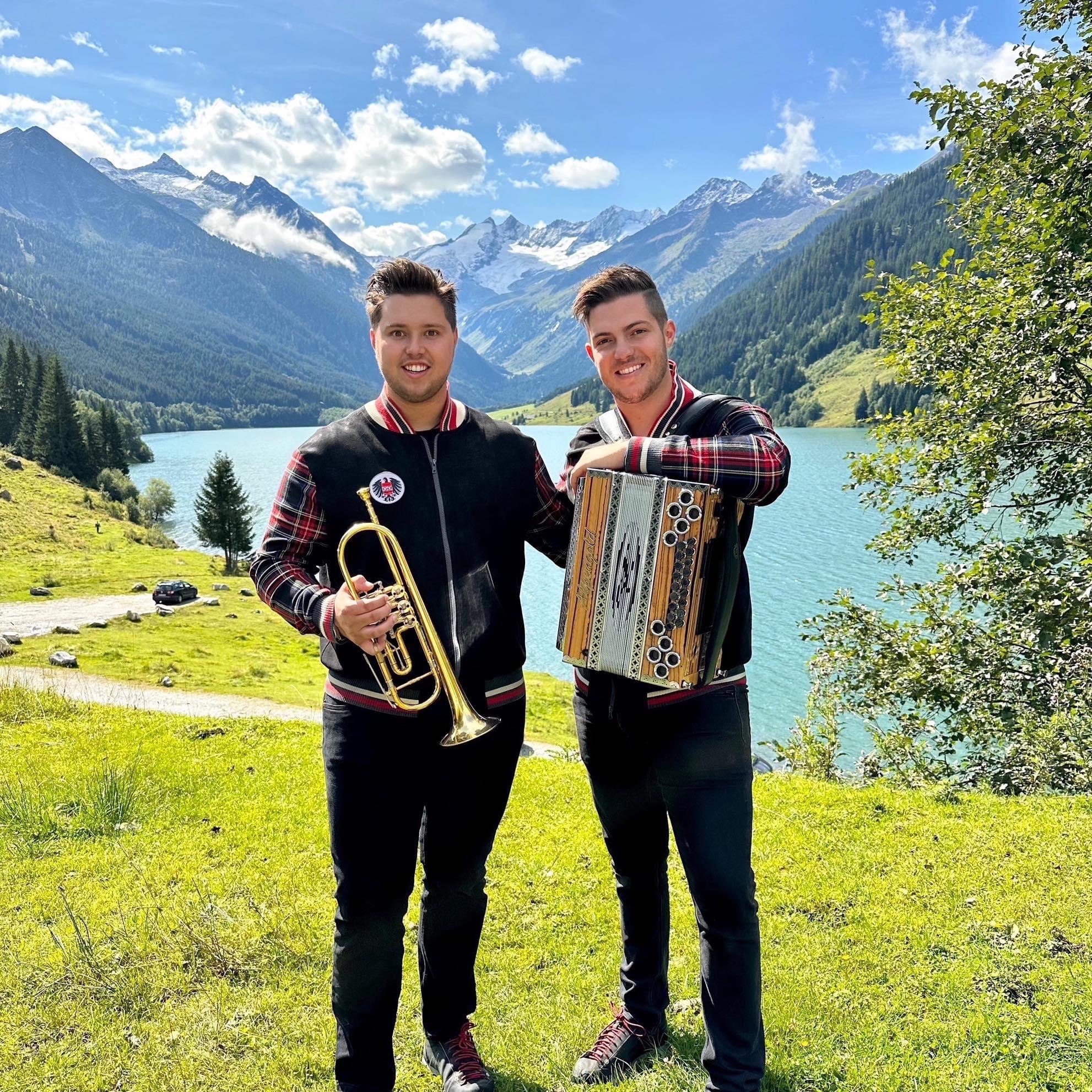 Zwei junge Männer mit Musikinstrumenten vor Bergsee und Bergen, sonniger Tag, grüne Wiese, Bäume im Hintergrund.
