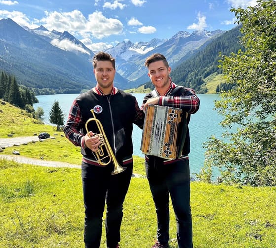 Two young men with instruments by a mountain lake, sunny day, green grass, mountains in the background.