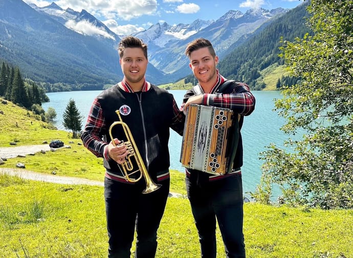 Two young men with instruments by a mountain lake, sunny day, green grass, mountains in the background.