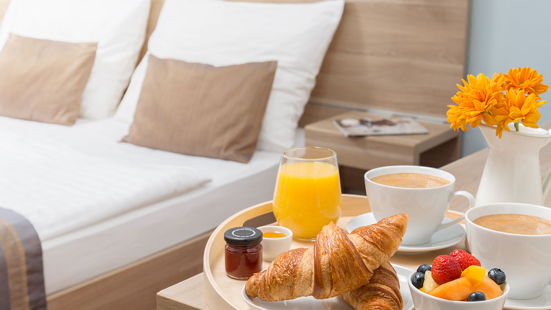 A tray with breakfast - croissants, fruit, coffee and orange juice - stands on a bedside table next to a cozy double bed with decorative pillows.