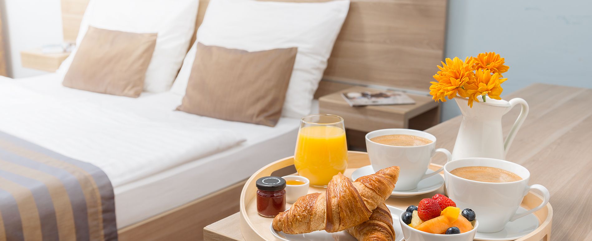 Ein Tablett mit Frühstück – Croissants, Obst, Kaffee und Orangensaft – steht auf einem Nachttisch neben einem gemütlich gemachten Doppelbett mit Zierkissen.
