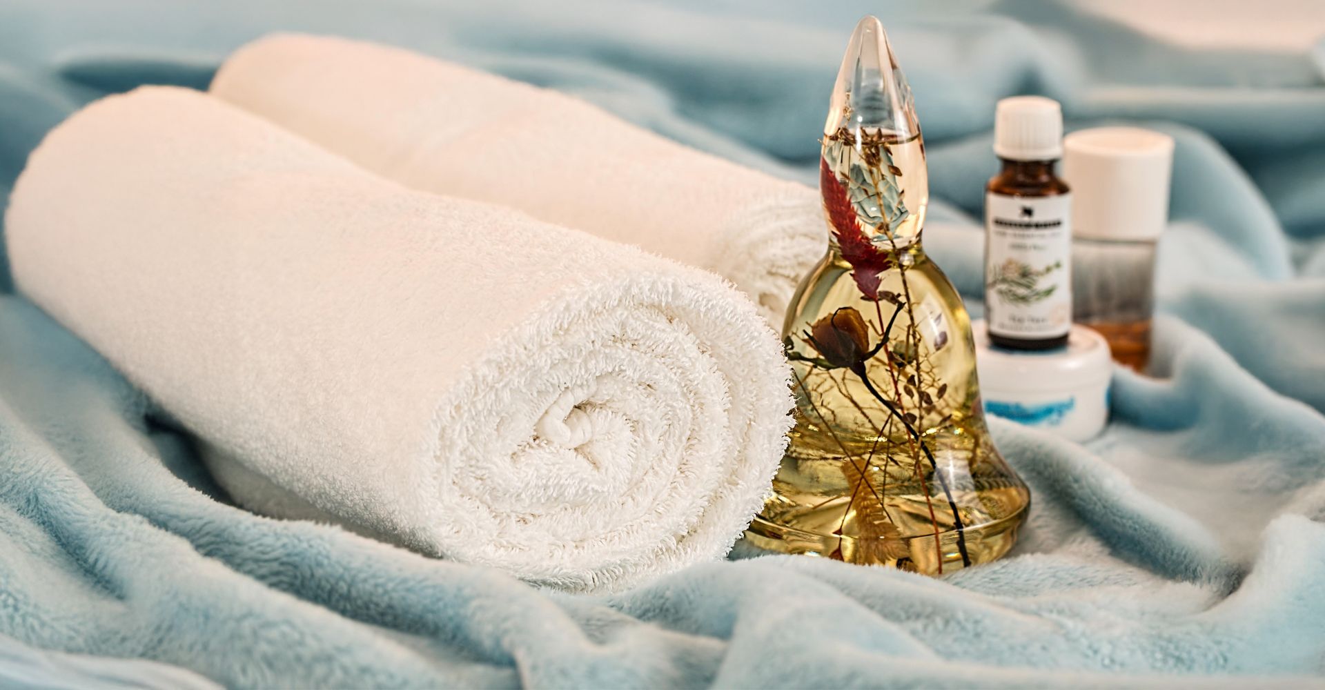 Two white towels, a golden Buddha figure, and small oil bottles rest on a soft blue cloth – a calming wellness setup.