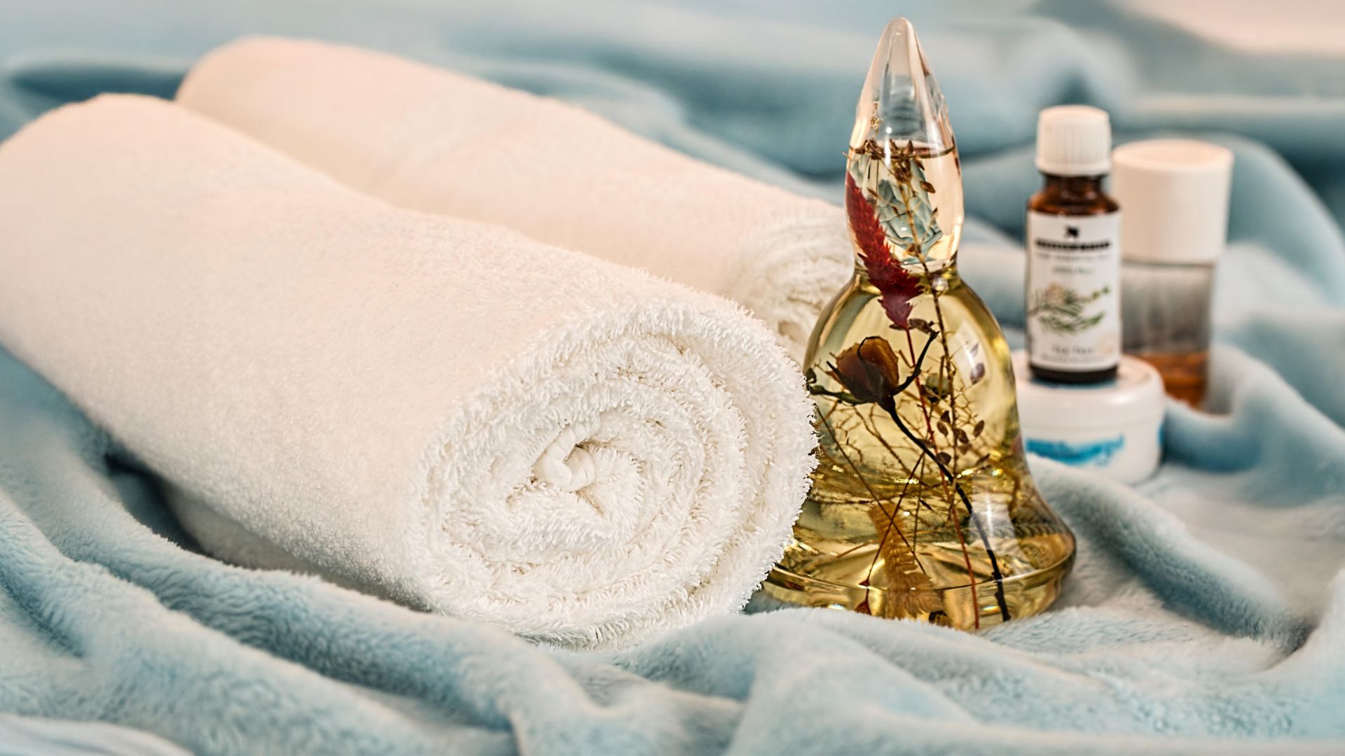 Zwei weiße Handtücher, eine goldene Buddha-Figur und kleine Fläschchen mit Öl stehen auf einem weichen, blauen Tuch – ein ruhiges Wellness-Arrangement.