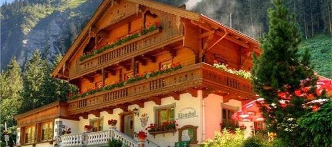 A multi-story chalet-style house with wooden facade, balcony, and flowering plants, surrounded by trees in the mountains.