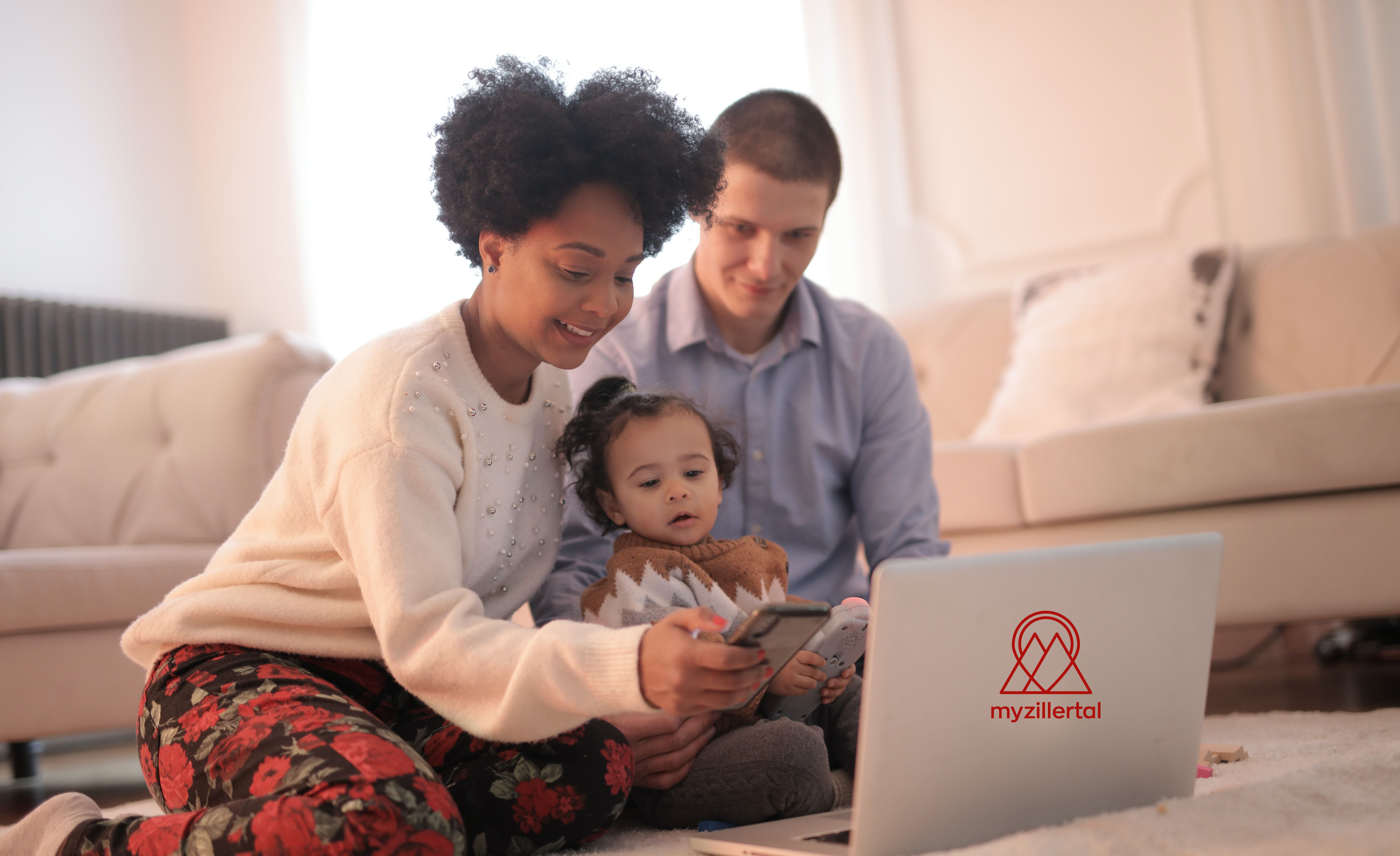 A young family sits on the floor in front of a laptop. The mother holds the baby, the father looks on with a smile. They look at the screen together. In the background is a sofa with cushions. The room is bright and feels cozy.