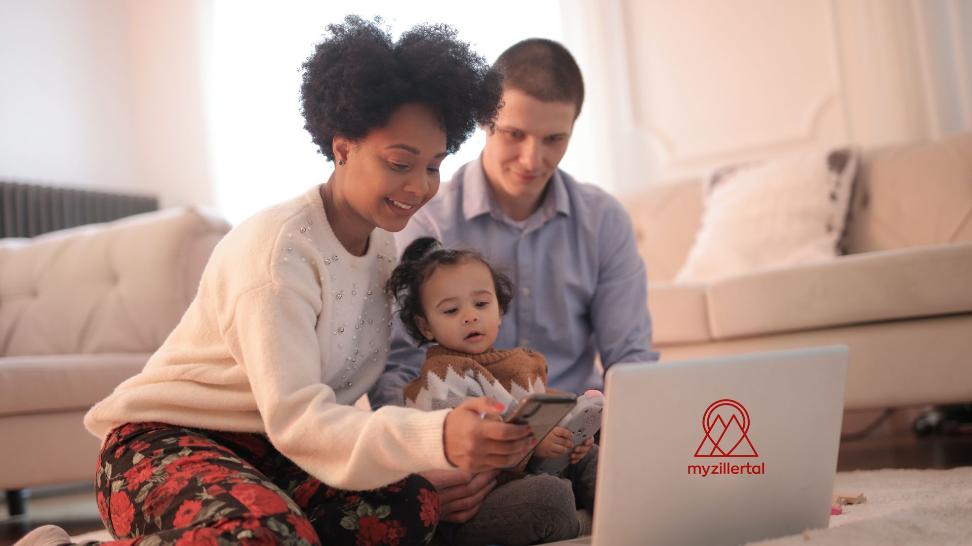 A young family sits on the floor in front of a laptop. The mother holds the baby, the father looks on with a smile. They look at the screen together. In the background is a sofa with cushions. The room is bright and feels cozy.