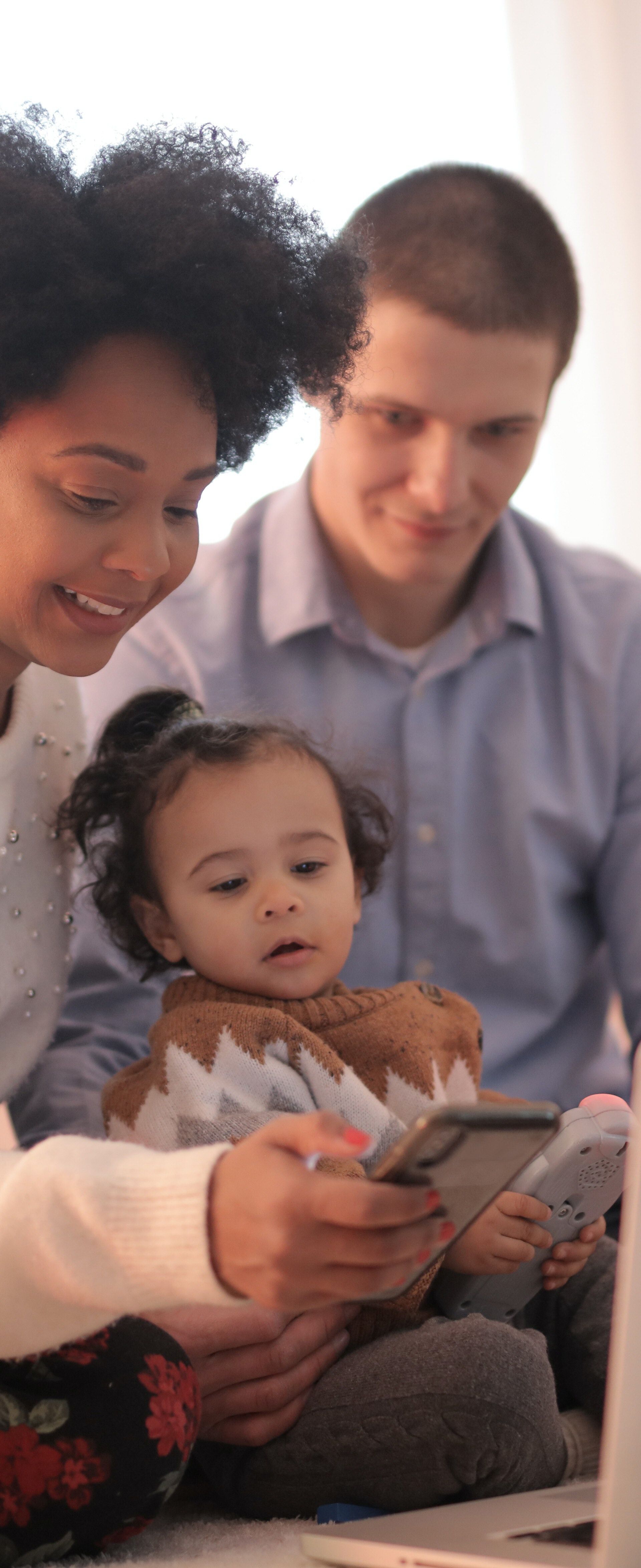 ine junge Familie sitzt auf dem Boden vor einem Laptop. Die Mutter hält das Baby, der Vater schaut lächelnd zu. Sie blicken gemeinsam auf den Bildschirm. Im Hintergrund steht ein Sofa mit Kissen. Der Raum ist hell und wirkt gemütlich.