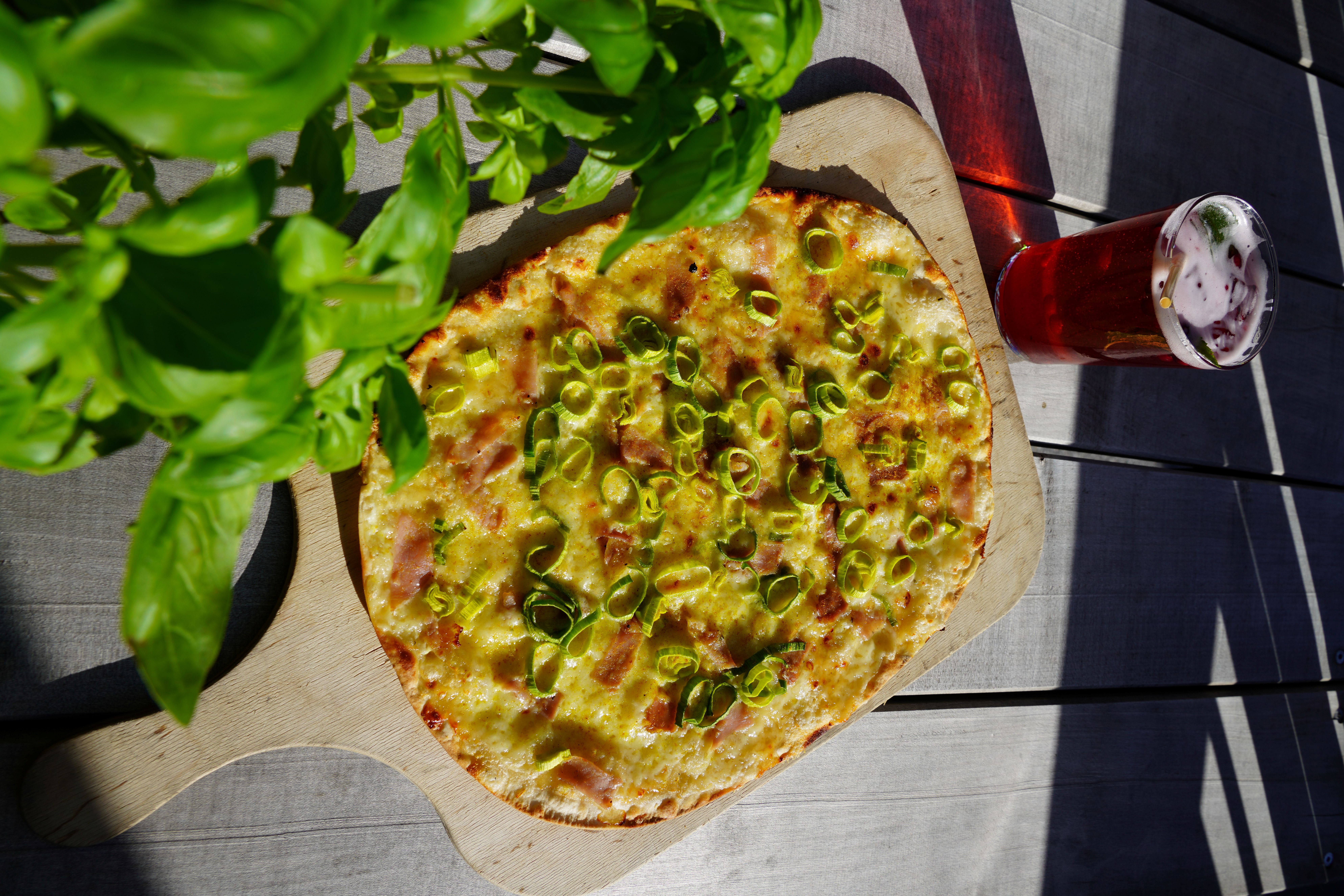 The image shows a crispy tarte flambée on a wooden board, topped with cheese, ham, and fresh leek. In the foreground, green basil leaves add a fresh touch to the image.